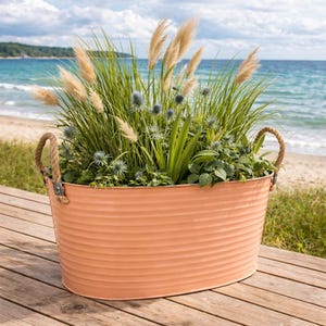 Op de afbeelding: Een ovale, perzikroze metalen plantenbak gevuld met hoog, groen gras, beige pluimen en blauwe distelachtige bloemen. De plantenbak heeft touwhandvatten en staat op een houten dek met een strand en oceaan op de achtergrond.