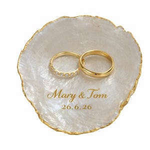 Puede incluir: Un plato nacarado con borde dorado sostiene dos anillos de boda dorados. Uno de los anillos está adornado con pequeñas gemas. El plato está inscrito con "Mary & Tom" y la fecha "26.6.26".
