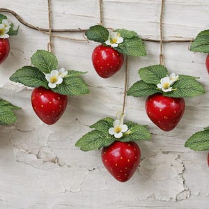 Könnte beinhalten: Künstliche Erdbeeren, die an Schnüren hängen, jede mit grünen Blättern und kleinen weißen Blüten. Die leuchtend roten Erdbeeren sind vor einem weißen, strukturierten Hintergrund angeordnet.