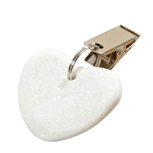 Könnte beinhalten: Ein weißer herzförmiger Clip mit einer silberfarbenen Metallklammer. Das Herz besteht aus einem strukturierten, möglicherweise steinartigen Material. Die Klammer ist mit einem kleinen Metallring am Herzen befestigt.