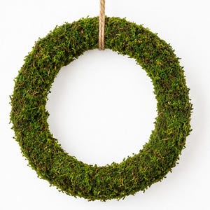Może przedstawiać: Okrągły wieniec pokryty gęstym, żywo zielonym mchem. Wieniec ma teksturowaną, organiczną powierzchnię i jest zawieszony na cienkiej, jasnobrązowej pętli ze sznurka jutowego przymocowanej u góry, tworząc prostą dekorację ścienną o naturalnym wyglądzie.
