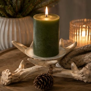 May include: A dark green pillar candle with a lit wick sits inside a white circular holder supported by a base shaped like light-colored deer antlers. A small brown pinecone is attached to the front of the antler base with twine.