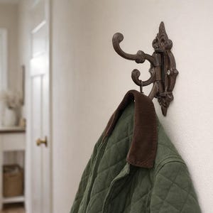 Può includere: Un gancio da parete in metallo marrone scuro e ornato con tre ganci, che tiene una giacca trapuntata verde con un colletto marrone. Il gancio è montato su una parete color crema, suggerendo un corridoio o un ingresso.