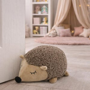 May include: A soft hedgehog doorstop in a child's bedroom. The doorstop is brown with a beige face, ears, and feet. It has closed eyes and a black nose. The room features a pink rug, a white bookshelf, and a canopy bed.
