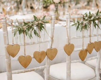 May include: Two white wooden chairs with heart-shaped wooden ornaments hanging from them. The chairs are decorated with greenery and are set up for a wedding or special event. The hearts are suspended by twine.