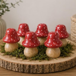 Op de afbeelding: Zes houten paddenstoelbeeldjes met rode doppen en witte stippen zijn gerangschikt op een houten schijf met groen mos. De paddenstoelen hebben een natuurlijke houten basis en zijn een decoratief item.