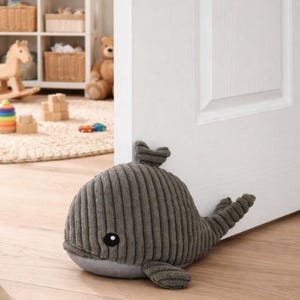 May include: A gray whale-shaped doorstop made of corduroy fabric. The doorstop has a rounded body, a small fin on top, and a tail. It is placed on a light-colored wooden floor next to a white door.