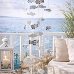 Può includere: Mobile decorativo con ornamenti a forma di pesce nei toni del bianco e blu. I pesci sono sospesi a un telaio di legno con fili, evocando un tema marittimo. Lo sfondo mostra il mare e il cielo.