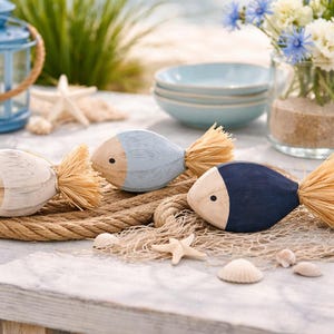 May include: Three decorative wooden fish with straw tails and two-tone bodies in blue, white, and navy. The fish rest on a rope and fishing net, with seashells and a blue lantern in the background. A coastal theme.