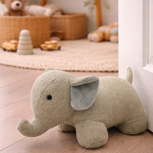 May include: A sage green fabric elephant doorstop with a textured surface, featuring a small trunk, a short tail, and soft grey inner ears. The elephant has a simple black bead eye and is positioned on a wooden floor next to a white door frame.