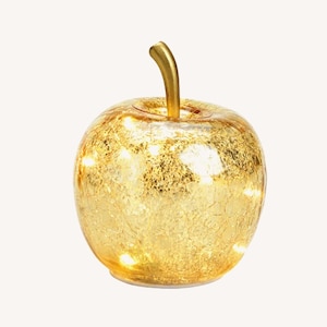 Puede incluir: Una lámpara decorativa con forma de manzana. La manzana de cristal está cubierta de pan de oro y tiene un tallo dorado. Pequeñas luces LED son visibles en el interior, creando un brillo cálido. Dimensiones: aproximadamente 10 cm de altura.