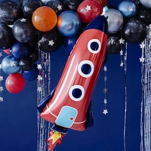 Puede incluir: Un globo rojo con forma de cohete con ventanas circulares blancas y detalles azules, rodeado de un grupo de globos de varios colores, incluyendo negro, azul, naranja y plateado. Guirnaldas de estrellas plateadas y un fondo azul oscuro completan la escena festiva.