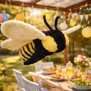 Puede incluir: Una decoración de abeja de peluche de gran tamaño, con rayas negras y amarillas, detalles amarillos esponjosos y grandes alas color crema. La abeja está suspendida en el aire, lo que sugiere una fiesta o un evento al aire libre. El fondo incluye decoraciones de fiesta y una mesa puesta.