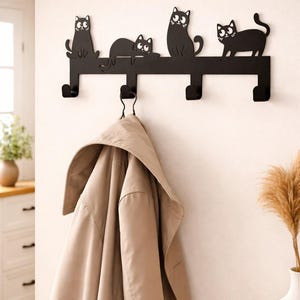 Könnte beinhalten: Schwarzer Metall-Wandgarderobenständer mit vier stilisierten schwarzen Katzen-Silhouetten. Der Ständer hat vier Haken und einen beigen Mantel, der an einem Haken hängt. Der Ständer ist an einer weißen Wand befestigt.