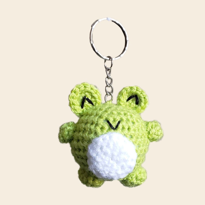 May include: A green crocheted frog keychain with a white belly and black eyes. The frog is attached to a silver key ring.