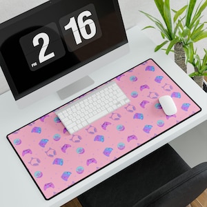 Puede incluir: Una alfombrilla de escritorio rectangular con un patrón repetitivo de mandos de juego, auriculares y consolas portátiles en tonos morados y azules sobre un fondo rosa suave. La alfombrilla está colocada sobre una superficie de escritorio blanca.