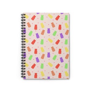 Op de afbeelding: Spiraalgebonden notitieboek met een herhalend patroon van kleurrijke gummyberen en kleine roze hartjes op een lichtroze achtergrond. Het notitieboek heeft een zwarte spiraalbinding.