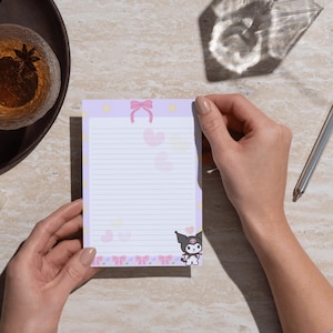 Puede incluir: Un bloc de notas rectangular con fondo morado claro, que presenta líneas horizontales para escribir. El diseño incluye un lazo rosa en la parte superior, estrellas amarillas a los lados, corazones pastel dispersos y una ilustración de Kuromi.