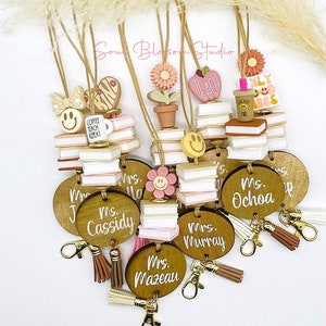 May include: A set of 8 wooden keychain charms with a teacher theme. Each charm features a round wooden tag with a teacher's name, a stack of books, and a small charm. The charms are decorated with pink, white, and brown colors. The charms are attached to gold key rings with brown tassels.