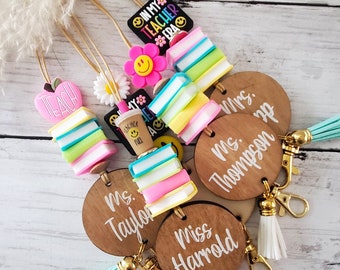 May include: Five wooden keychains with colorful book stacks, a teacher apple, and a smiley face charm. Each keychain has a different teacher's name: Ms. Taylor, Miss Harrold, Mrs. Thompson, Ms. p, and a keychain with the text "TEACH".