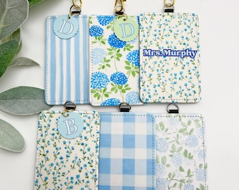 May include: Six rectangular ID badge holders featuring various blue and white patterns including stripes, gingham, and floral prints. Each holder is attached to a lanyard with wooden, white, and blue beads. Some holders feature circular letter charms or the text Mrs. Murphy.