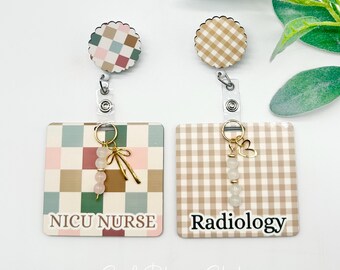 May include: Two square badge reels with scalloped circular tops. The left reel features a multicolored checkered pattern with the text NICU NURSE and a charm with three beads and a bow. The right reel has a tan gingham pattern, the text Radiology, and a charm with three beads and a heart.