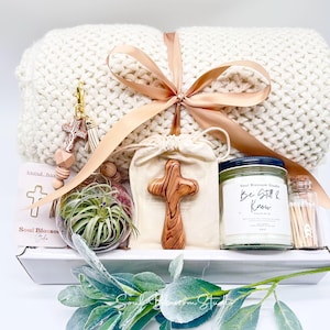 May include: A gift set featuring a cream-colored knitted blanket tied with a peach ribbon. The set includes a wooden cross, a candle, a small air plant, and a cross-shaped keychain. The candle jar has a black lid and a label that reads "Be Still & Know."
