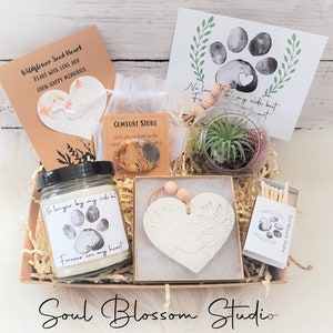 May include: A gift box with a variety of items, including a candle with a paw print design, a heart-shaped ornament, a terrarium, and a card with a paw print. The box also contains a comfort stone and a matchbox. The text "Soul Blossom Studio" is visible.