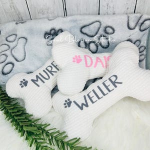 May include: Three white plush dog bone toys with paw prints and names "Muf", "Dax", and "Weller" embroidered on them. The toys are on a white fluffy surface with a gray and white paw print blanket in the background.