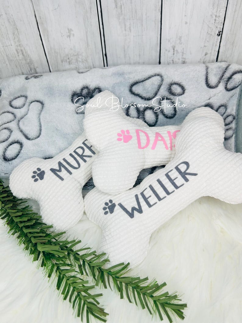 May include: Three white plush dog bone toys with paw prints and names "Muf", "Dax", and "Weller" embroidered on them. The toys are on a white fluffy surface with a gray and white paw print blanket in the background.