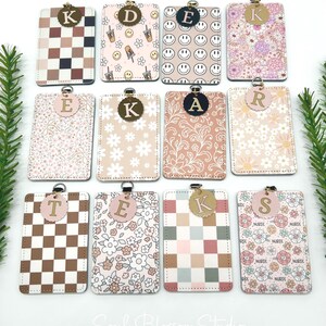 May include: Twelve rectangular badge holders with rounded corners, each featuring a unique pattern including checkered, floral, and smiley face designs. Each holder has a metal ring at the top and a circular tag displaying a single letter.