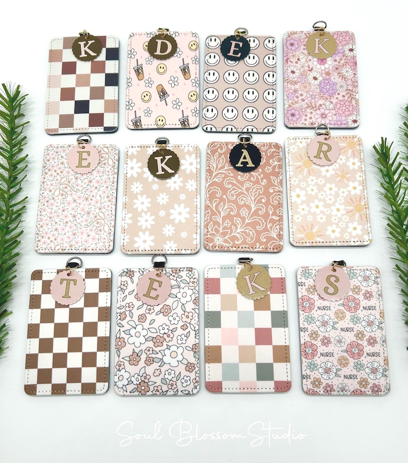 May include: Twelve rectangular badge holders with rounded corners, each featuring a unique pattern including checkered, floral, and smiley face designs. Each holder has a metal ring at the top and a circular tag displaying a single letter.
