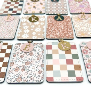 May include: A collection of rectangular ID badge holders featuring various patterns including brown and white checkers, floral prints, and smiley faces. Each holder is topped with a circular charm displaying a single gold letter, such as T, E, K, A, R, or S.