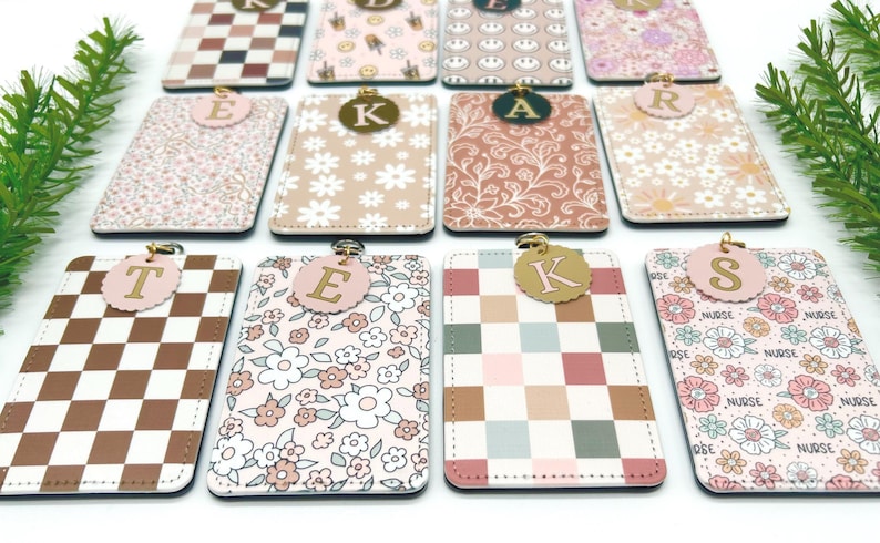 May include: A collection of rectangular ID badge holders featuring various patterns including brown and white checkers, floral prints, and smiley faces. Each holder is topped with a circular charm displaying a single gold letter, such as T, E, K, A, R, or S.