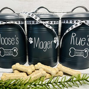 May include: Three black metal canisters with white lids and white ribbon accents. Each canister has a white paw print and a white bone with the text "treats" on it. The canisters are labeled "Moose's Treats", "Mager's Treats", and "Rue's Treats".