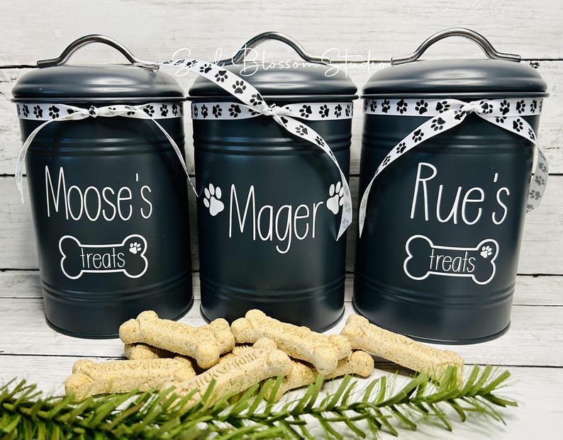May include: Three black metal canisters with white lids and white ribbon accents. Each canister has a white paw print and a white bone with the text "treats" on it. The canisters are labeled "Moose's Treats", "Mager's Treats", and "Rue's Treats".