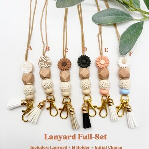 May include: Six lanyards labeled A through F, each featuring a gold-toned cord, a wooden bead, a textured cream bead, a silicone flower or patterned disc, and a gold-toned lobster clasp with a tassel. Text reads: Lanyard Full-Set Includes: Lanyard + Id Holder + Initial Charm.