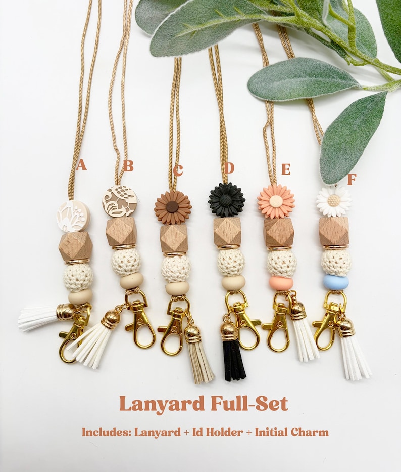 May include: Six lanyards labeled A through F, each featuring a gold-toned cord, a wooden bead, a textured cream bead, a silicone flower or patterned disc, and a gold-toned lobster clasp with a tassel. Text reads: Lanyard Full-Set Includes: Lanyard + Id Holder + Initial Charm.