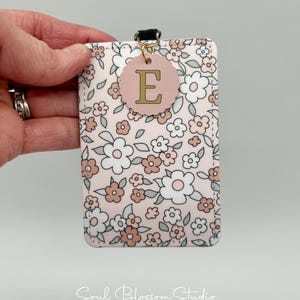 May include: A rectangular ID badge holder featuring a floral pattern with white, light pink, and sage green flowers on a pale pink background. A scalloped circular tag with the letter E in gold is attached to the top with a gold jump ring.
