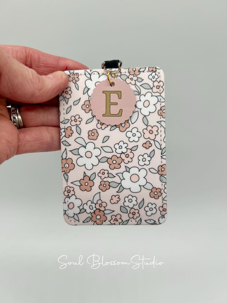 May include: A rectangular ID badge holder featuring a floral pattern with white, light pink, and sage green flowers on a pale pink background. A scalloped circular tag with the letter E in gold is attached to the top with a gold jump ring.