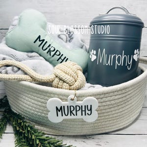 May include: A white wicker basket filled with dog toys, including a green bone-shaped toy, a rope toy with a knotted end, and a white bone-shaped tag with the name "Murphy" on it. A gray metal food container with the name "Murphy" and paw prints is also in the basket.