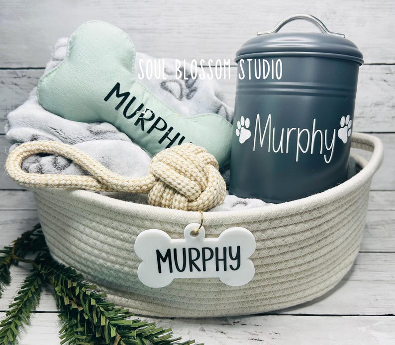 May include: A white wicker basket filled with dog toys, including a green bone-shaped toy, a rope toy with a knotted end, and a white bone-shaped tag with the name "Murphy" on it. A gray metal food container with the name "Murphy" and paw prints is also in the basket.