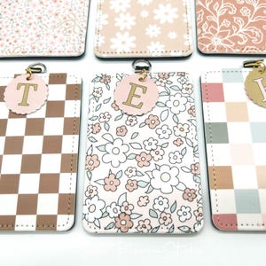 May include: A collection of rectangular ID badge holders featuring various patterns including brown and white checkers, floral prints, and multi-colored checkered designs. Each holder includes a circular tag with a letter T, E, or K attached.