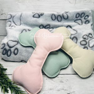 May include: Three plush dog bone toys in pink, green, and cream colors. The toys are lying on a gray and white blanket with paw prints.
