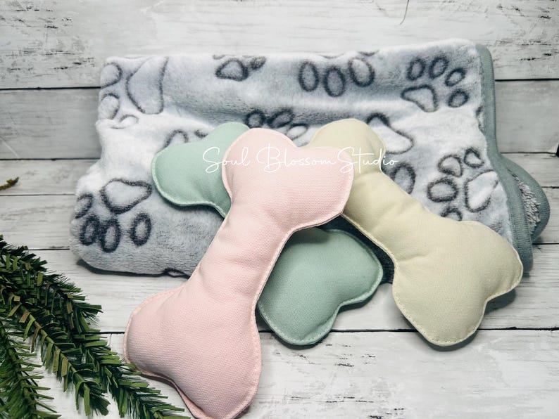 May include: Three plush dog bone toys in pink, green, and cream colors. The toys are lying on a gray and white blanket with paw prints.