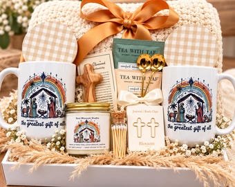 May include: A gift set featuring two white mugs with a nativity scene and rainbow graphic, a candle, a wooden cross, two gold spoons, tea packets, and cross-shaped paper clips. The mugs are printed with the text, the greatest gift of all.