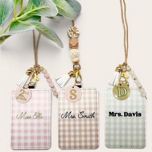 May include: Three rectangular ID badge holders with gingham patterns in pink, tan, and light green. Each holder features a gold-toned lanyard, a white tassel, a beaded accent, and a circular charm with the text 2nd grade, S, or D, and names.
