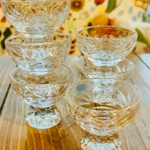May include: A set of six clear, stemmed dessert bowls. Each bowl features a detailed, cut-glass design with a floral pattern. The bowls are arranged on a wooden surface, showcasing their intricate craftsmanship and elegant style. Ideal for serving small portions.