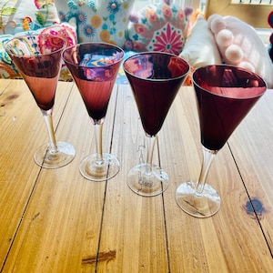 May include: Four stemmed wine glasses with a deep red bowl and clear stem and base. The glasses are arranged on a wooden surface. The glasses have a reflective quality, and the light catches the inside of the bowls.