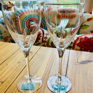 May include: Two clear crystal wine glasses with gold rims. The glasses have a delicate, etched design and sit on a wooden table. Each glass has a small, rectangular tag at the base. The background includes colorful pillows and a window.